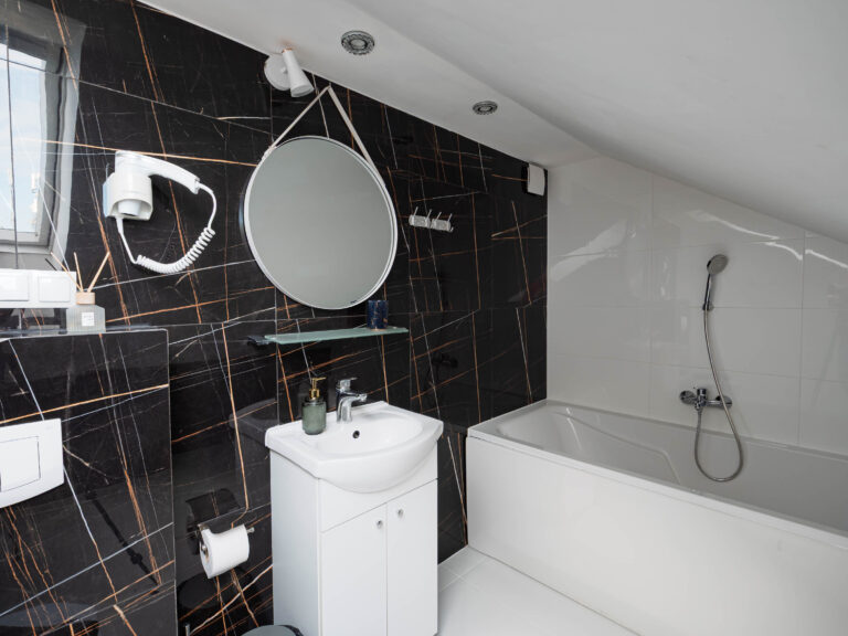 Elegancka łazienka apartamentu z umywalką, okrągłym lustrem, wanną i czarnymi płytkami